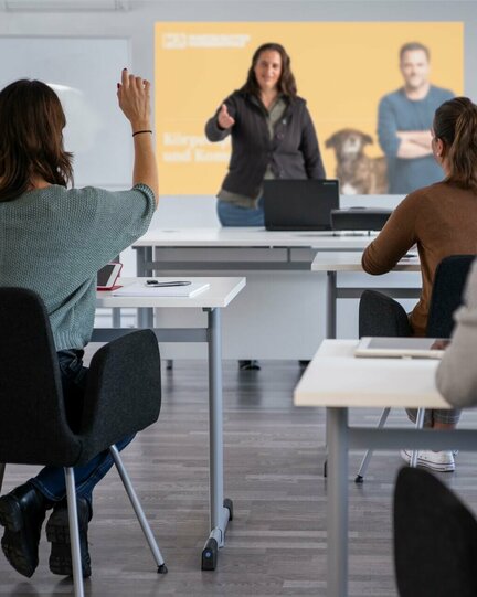 Klassenraum mit mehreren Tischen und Stühlen, die von Menschen besetzt sind