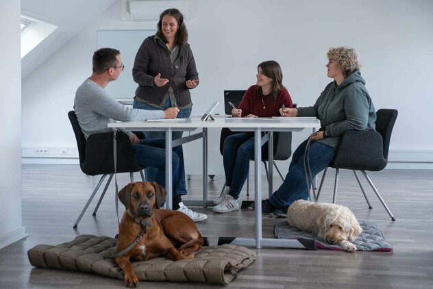 Ein großer Tisch, an dem drei Personen sitzen und eine vierte Person steht