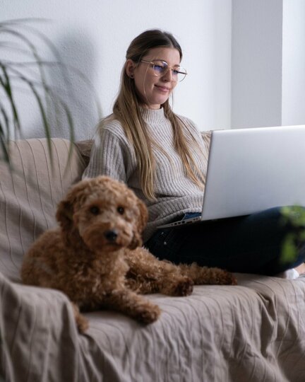 Frau sitzt mit ihrem Hund auf dem Sofa und hat ihren Laptop auf dem Schoß